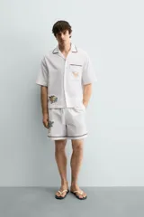 Bermuda de corte relajado confeccionada en algodón, color blanco con vivo en contraste color marrón en el ruedo. Presenta cintura elástica con cordón ajustable y un bordado decorativo en la pierna derecha que incluye una palmera y un gallo.