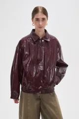 Chaqueta de eco-cuero en color bordó con acabado brillante, diseño holgado, cuello camisero, cierre frontal con botones a presión y detalle de elástico fruncido en el ruedo.