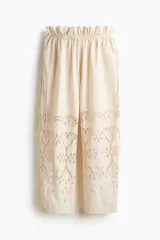 Pantalón oversized color beige, con cintura paper bag ajustable con cordones a los lados y piernas de corte barrel con detalles de broderie en la parte inferior.