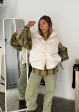 Chaleco color blanco hueso de peluche con cuello alto, cierre frontal con botones a presión y detalles en cuero sintético color beige.