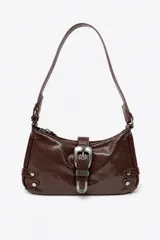 Bolso de hombro de poliuretano efecto charol arrugado en color marrón, con cierre de cremallera superior. Cuenta con una hebilla decorativa grande de metal plateado envejecido en el frente, tiras laterales con tachas metálicas y asa ajustable.