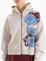 Buzo con cierre frontal y capucha, color beige, con estampado floral y de cadenas en el frente. La capucha cuenta con forro de corderito al tono, mangas largas con puños elásticos y ajuste holgado.
