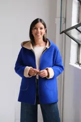 Tapado de paño color azul con capucha, cierre frontal y detalles en color beige en el interior de la capucha, puños y bordes de los bolsillos.