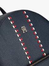 Mochila de diseño minimalista con acabado granulado en color azul marino. Presenta un bolsillo frontal con cierre, correas ajustables, asa superior y el monograma TH metálico en la parte frontal. Se destaca por una cinta decorativa vertical con detalles en rojo y blanco.