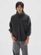 Buzo de sherpa color gris oscuro, con cuello alto y medio cierre frontal. Presenta un bolsillo ojal en el pecho con cierre y bolsillos laterales. Cuenta con cordón regulable en el ruedo para un ajuste personalizado.