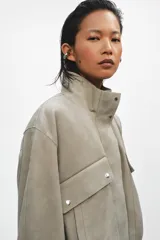 Chaqueta corta de ante con cuello alto, manga larga y cierre frontal con cremallera y botones a presión. Presenta bolsillos frontales con solapa y bajo abullonado con pliegues.