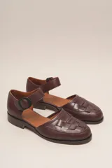 Zapato de cuero color borgoña, con puntera redondeada y diseño trenzado. Presenta una tira sobre el empeine con hebilla metálica negra.
