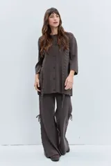 Cardigan de punto color marrón, con cierre frontal de botones, cuello redondo y mangas tres cuartos. Presenta un diseño con detalles de cordones entrelazados en los laterales que se extienden hacia el ruedo.