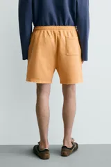 Bermuda de corte relajado confeccionada en tejido de algodón con interior de rizo. Presenta cintura elástica con cordón ajustable, bolsillos laterales, un bolsillo trasero de plastrón y bajo con acabado irregular con efecto lavado.