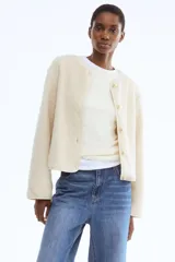 Chaqueta de sherpa suave con cuello redondo, corte holgado y cierre frontal mediante botones metálicos dorados. Presenta ribetes de cinta fina en los bordes y mangas largas.