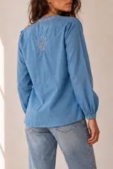 Blusa blanca de manga larga con detalles bordados en tono marrón en el pecho y las mangas, cuello redondo y cierre frontal con pequeños botones.
