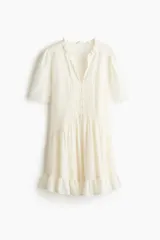 Vestido túnica corto color blanco marfil, con escote en V y cuello de volados. Presenta mangas cortas abullonadas, botonadura frontal parcial y detalles de alforzas e inserciones de encaje en el frente. Cuenta con costuras fruncidas en la cadera y ribete de volado en el bajo.