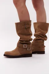 Bota de caña alta color marrón chocolate, confeccionada en gamuza. Presenta dos correas desmontables con hebillas metálicas plateadas.