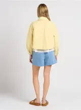 Chaqueta casual color amarillo pastel, de corte corto y suelto. Tiene cuello camisero, cierre frontal con botones metálicos, mangas largas y grandes bolsillos de parche en la parte delantera.