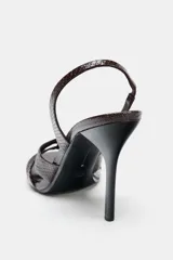 Sandalia de tacón alto fino, color marrón oscuro con textura de relieve de piel de serpiente. Presenta tiras anchas cruzadas en la parte delantera y una tira trasera con elástico ajustable. La punta es cuadrada.