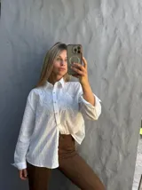 Camisa blanca de manga larga con diseño de rayas verticales sutiles y bordado floral en la parte superior delantera. Presenta cuello clásico y cierre frontal con botones.