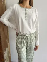 Pijama de dos piezas compuesto por una remera de manga larga con cuello redondo y cierre frontal parcial con botones, y un pantalón de corte holgado con cintura elástica y cordón ajustable. La remera es de color blanco con detalles en verde, mientras que el pantalón presenta un diseño a rayas horizontales blancas y verdes.