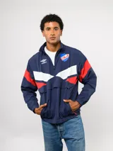 Buzo de nylon con medio cierre, diseño deportivo con bloques de color azul, blanco y rojo. Presenta el logo de Umbro y el escudo del Club Nacional de Football en el frente.