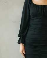 Vestido corto negro con escote recto, mangas largas abullonadas y diseño ajustado con fruncido en toda la parte frontal. Presenta un detalle de volado en el ruedo.