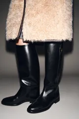 Bota de caña alta color negro, estilo hípico, con punta redonda, suela baja y tira decorativa con hebilla dorada en la caña.