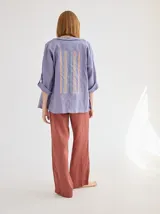 Chaqueta de lino color azul lavanda, de corte holgado, con escote redondo, cierre frontal con botones y mangas ajustables. Presenta un diseño versátil con bordado en la espalda.