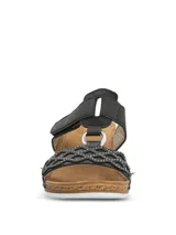 Sandalia Hush Puppies de plataforma baja con diseño de tiras negras. La tira delantera presenta un patrón geométrico calado con pequeñas tachas metálicas, mientras que la tira superior cuenta con un cierre de velcro para un ajuste personalizado. La plantilla es de color marrón y la suela tiene un acabado texturizado que simula madera.