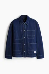 Sobrecamisa de denim de algodón rígido, color azul, con cuello inglés, bolsillos de parche en el pecho y en la parte inferior, y cierre frontal de botones metálicos.