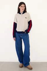 Sweater de punto acanalado con cuello alto y cierre frontal parcial. Presenta un diseño tricolor en bloques con cuerpo color crema, cuello y puños azul marino, y detalles en rojo en las mangas. Incluye una pequeña etiqueta con logo en el pecho.