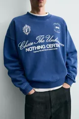 Sudadera de corte boxy fit en color azul con efecto lavado, cuello redondo y manga larga. Presenta estampado frontal con tipografía variada y gráficos estilo universitario. Interior afelpado y acabados en rib.