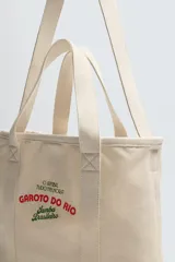 Bolso tipo shopper color crudo, fabricado en tejido de algodón con estampado de texto en verde y rojo. Tiene compartimento principal con cierre mediante imán, bolsillo interior con cierre y otro plano. Posee dos asas de mano.