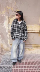 Camisa de manga larga con corte boxy fit, confeccionada en algodón a cuadros en tonos negro y gris. Presenta un bolsillo frontal en el pecho y el texto 'posta.' estampado en color blanco en la parte delantera.