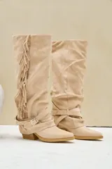Bota texana alta de gamuza color beige, con taco cuadrado, flecos largos colgantes en el lateral y tiras con hebilla metálica alrededor del tobillo.