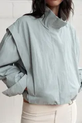 Chaqueta corta de cuello subido con manga larga, cierre frontal con cremallera y botones a presión ocultos. Presenta bolsillos delanteros y puños con pliegues.