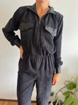 Mono largo de pana color negro, con cuello camisero y cierre frontal con cremallera. Presenta bolsillos con solapa de cuero en el pecho y puños de cuero.