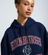Buzo tipo canguro de corte oversized, color azul marino, con capucha ajustable y bolsillo frontal. Presenta un estampado estilo college en el pecho con la inscripción 'Edinburgh Scotland' en letras rojas y blancas.