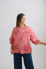 Blusa color coral de corte holgado, con cuello mao y cierre frontal de botones. Presenta mangas largas abullonadas y detalles de flores bordadas con técnica de calado en el frente.