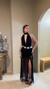 Vestido largo negro con escote profundo tipo halter y cuello alto. La falda presenta frunces y capas de microtul transparente. Se complementa con un cinturón ancho metálico de doble hebilla.