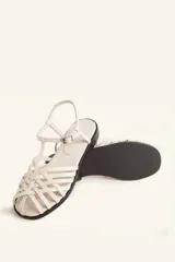 Sandalia plana de cuero blanco con diseño de tiras trenzadas en la parte delantera. Cuenta con una tira ajustable al tobillo con hebilla metálica y suela plana de color negro.