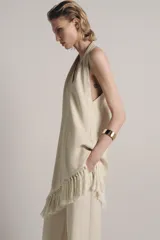 Top halter color crudo, de escote en V y diseño confeccionado en hilatura con mezcla de viscosa. Presenta bajo asimétrico con flecos al tono y cierre en la espalda.