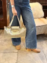 Cartera tipo baguette de cuero con acabado metalizado dorado, diseño rectangular con solapa frontal y cierre metálico a presión. Cuenta con doble asa fija para llevar al hombro.