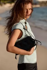 Bolso de hombro de material efecto acharolado negro, con doble compartimento exterior, cierre de cremallera y detalles de lazos en los laterales.