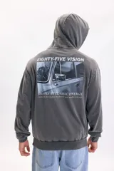 Hoodie de color gris oscuro con efecto lavado, capucha y bolsillo tipo canguro. Presenta una gran estampa en la espalda con la inscripción 'Eighty-Five Vision', una fotografía en blanco y negro de un automóvil clásico y texto descriptivo.