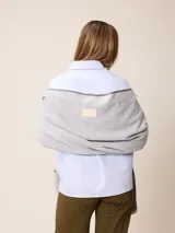 Poncho abierto de tejido de punto en color gris claro con bordes y detalles de bolsillos en gris oscuro. Presenta un diseño holgado y versátil con placa metálica decorativa en uno de los bolsillos frontales.