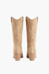 Bota de caña media color beige, confeccionada en suede, con pespuntes decorativos en la caña y la puntera. Presenta un tacón de bloque y punta en pico.
