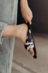 Sandalia tipo flip-flop con estampa cow print en blanco y negro. Presenta una tira ancha acolchonada que abraza el pie y una base negra lisa. Diseño liviano y cómodo de la marca Anne Chaussure.
