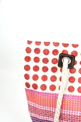 Bolso tipo tote con diseño de paneles en bloques que combinan estampado de lunares rojos sobre fondo blanco y franjas horizontales en tonos rosados, naranjas y morados. Cuenta con asas de soga gruesa de algodón en color crudo, sujetas con ojales metálicos y refuerzos de cuerina marrón.
