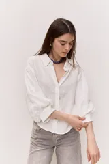 Camisa blanca de corte amplio y largo cropped, con mangas abullonadas, puño ajustado, cuello clásico y abertura en V con botones al tono.