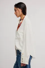 Blusa de lino color blanco crudo, de corte relajado, con cuello clásico, cierre frontal de botones, bolsillo en el pecho y mangas largas con presillas para arremangar.