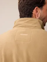 Campera de polar color beige con cierre frontal completo y cuello alto. Presenta un bolsillo en el pecho con cierre y detalle de parche con logo. El diseño combina paneles de tejido polar con detalles en tejido técnico en la zona del pecho.
