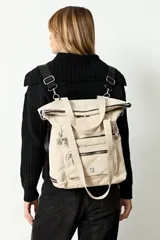 Mochila shopper color beige, confeccionada en poliéster. Presenta un diseño versátil con múltiples cierres, compartimento inferior expandible y un bolsillo frontal removible que funciona como estuche. Incluye correas ajustables y removibles para usar como mochila o bandolera, además de asas fijas para llevar al hombro.